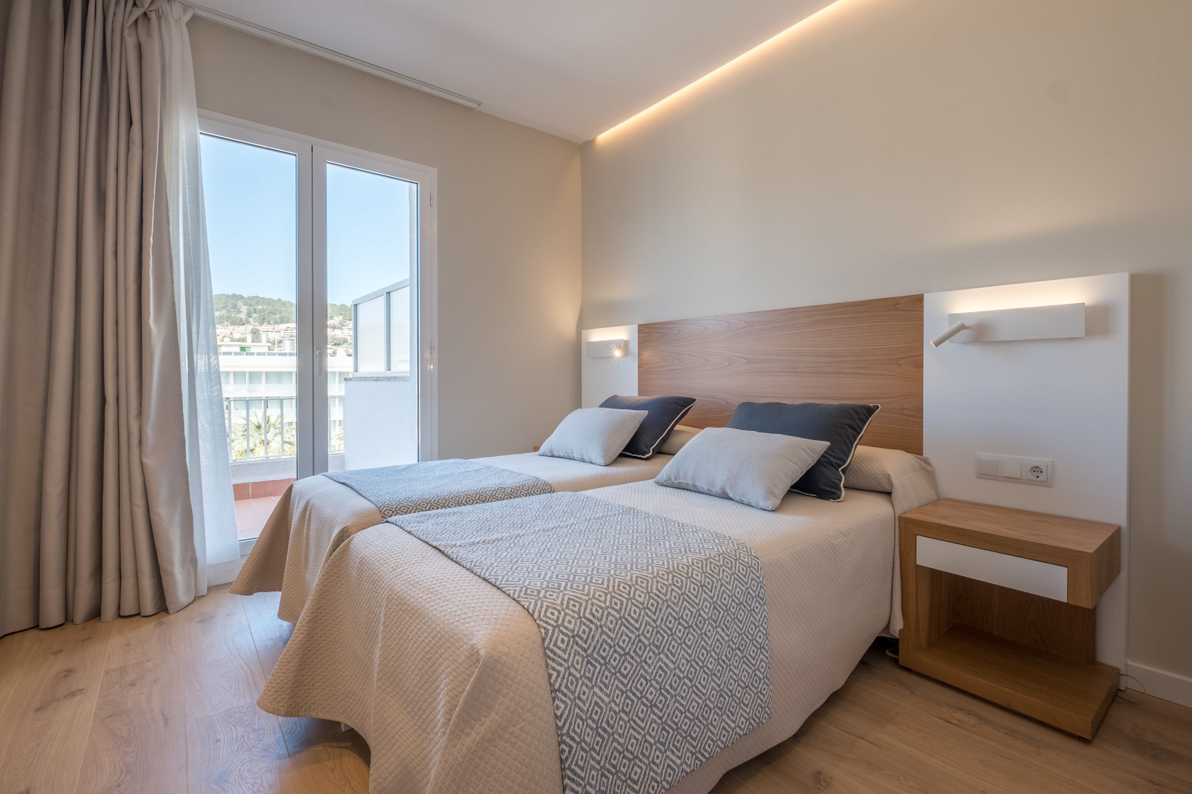 A modern hotel room with two beds, a wooden headboard, bedside tables, and a balcony with a view.