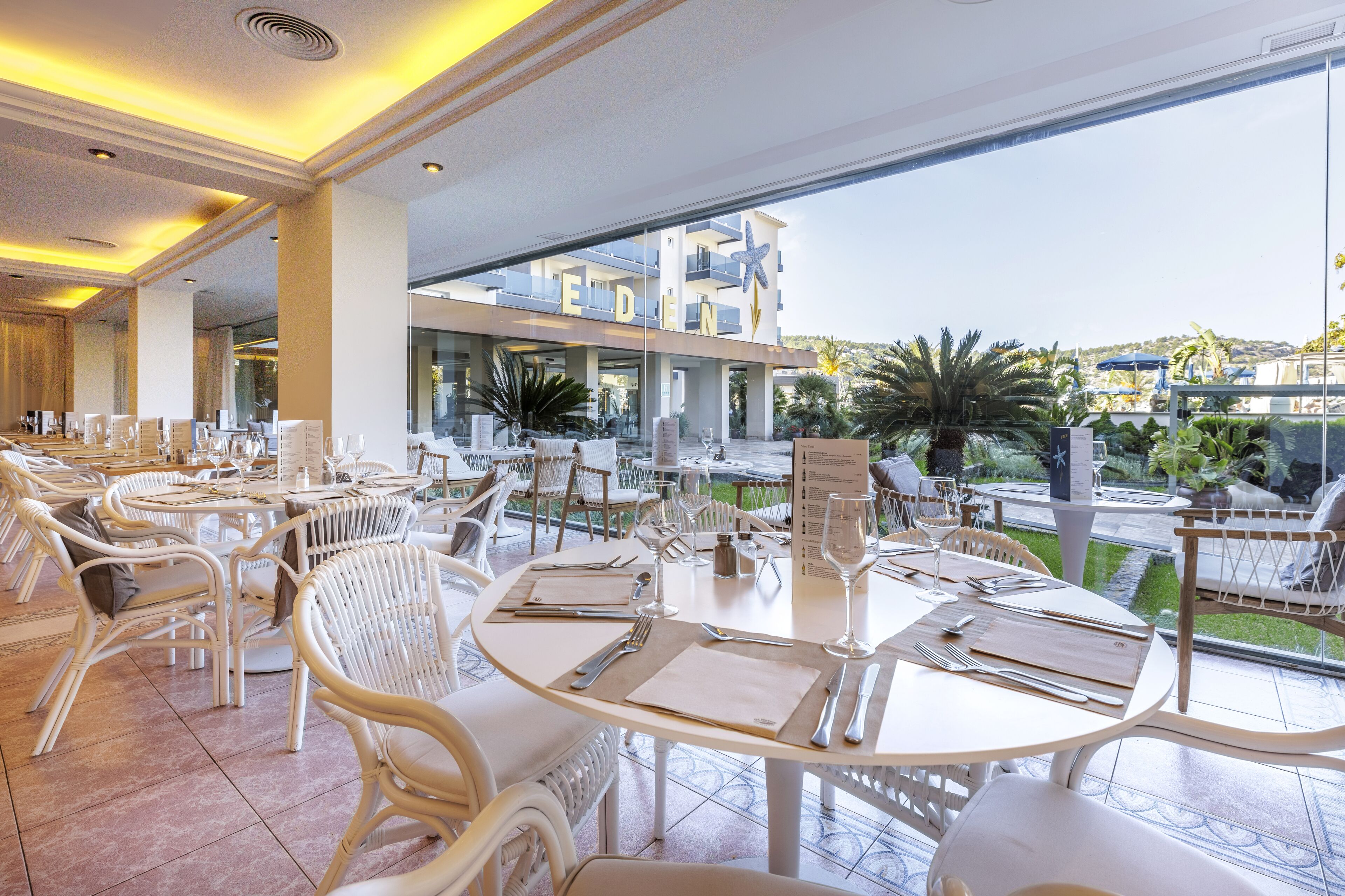 A restaurant with white wicker chairs and tables, a view of a pool and greenery, and a sign with the word "EDEN".