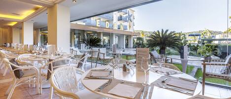 A restaurant with white wicker chairs and tables, a view of a pool and greenery, and a sign with the word "EDEN".