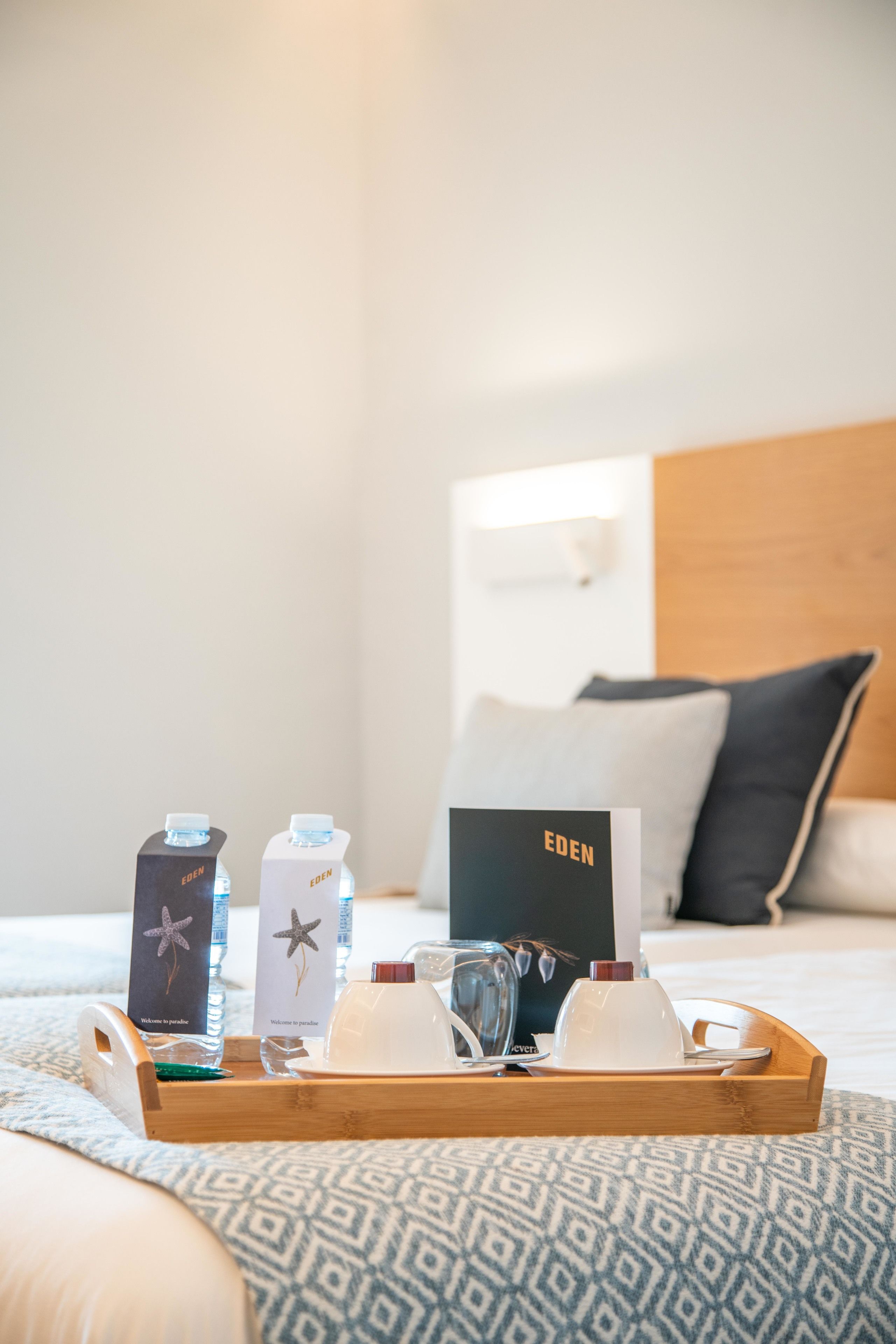 A neatly arranged hotel room with a bed, a wooden headboard, a tray with a welcome card, water bottles, a glass, and a cup.
