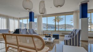 A spacious living area with striped sofas, a rattan chair, and a white coffee table. Large windows offer a view of the sea and hills.