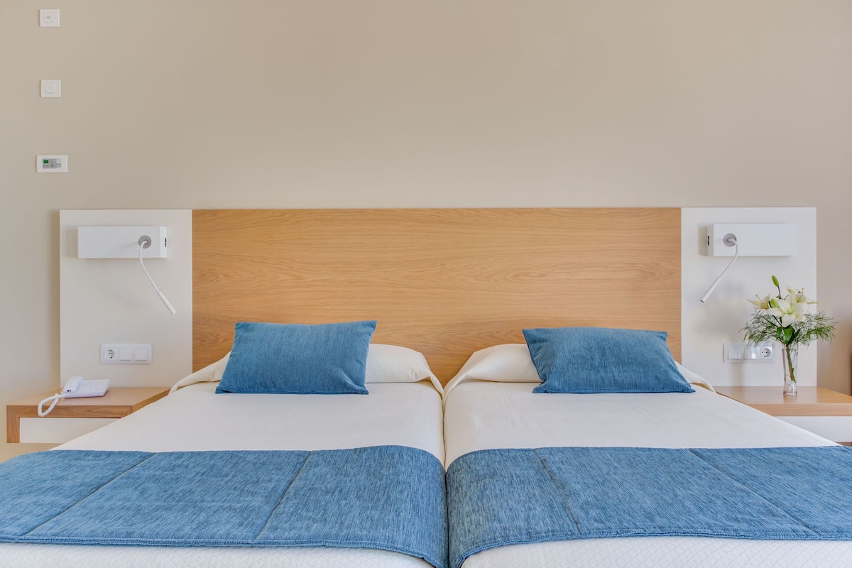 A double bed with blue bedding, a wooden headboard, bedside tables with lamps, and a small vase with flowers.