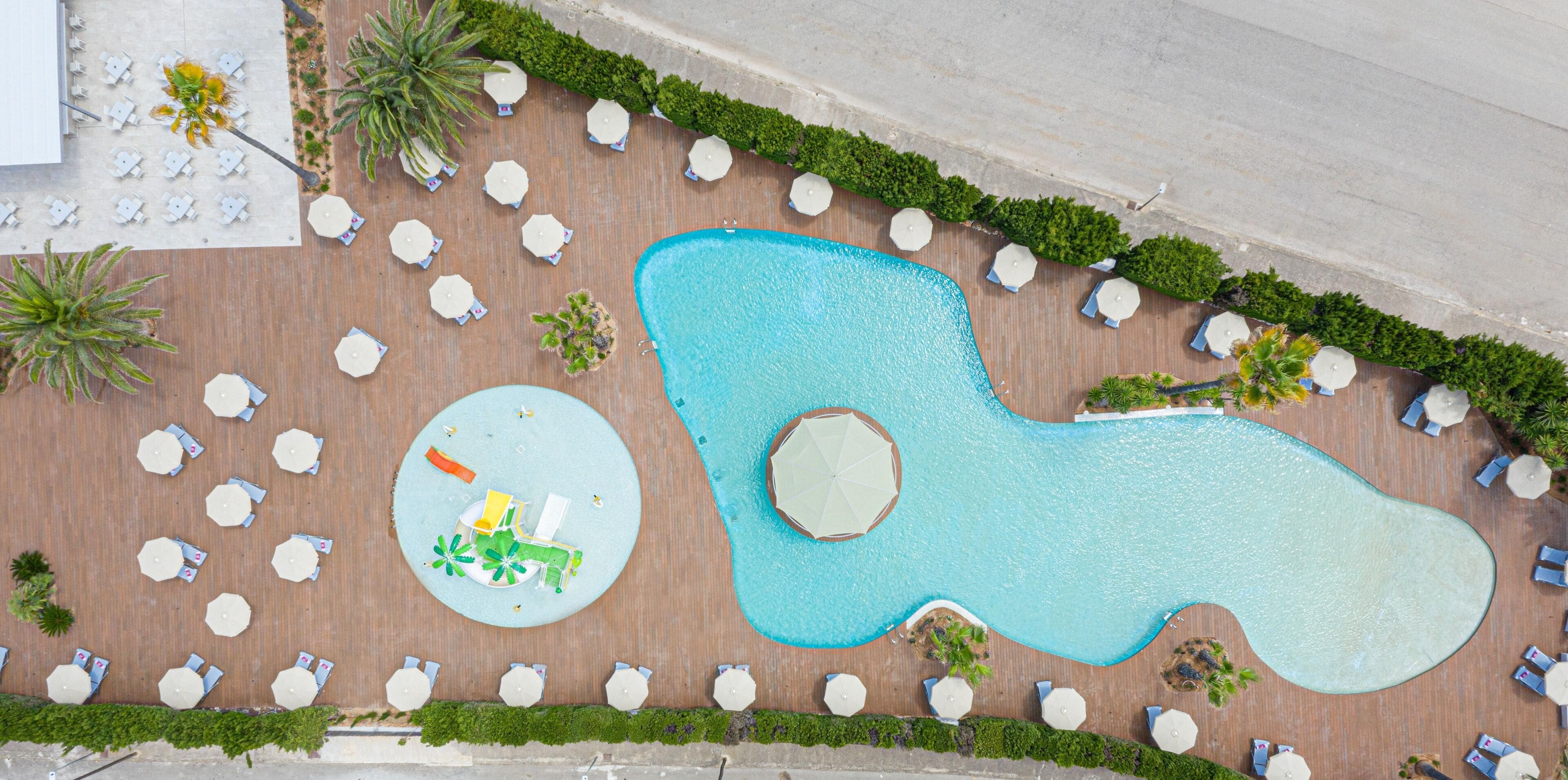 An aerial view of a pool area with multiple lounge chairs and umbrellas, surrounded by greenery and a unique pool shape.