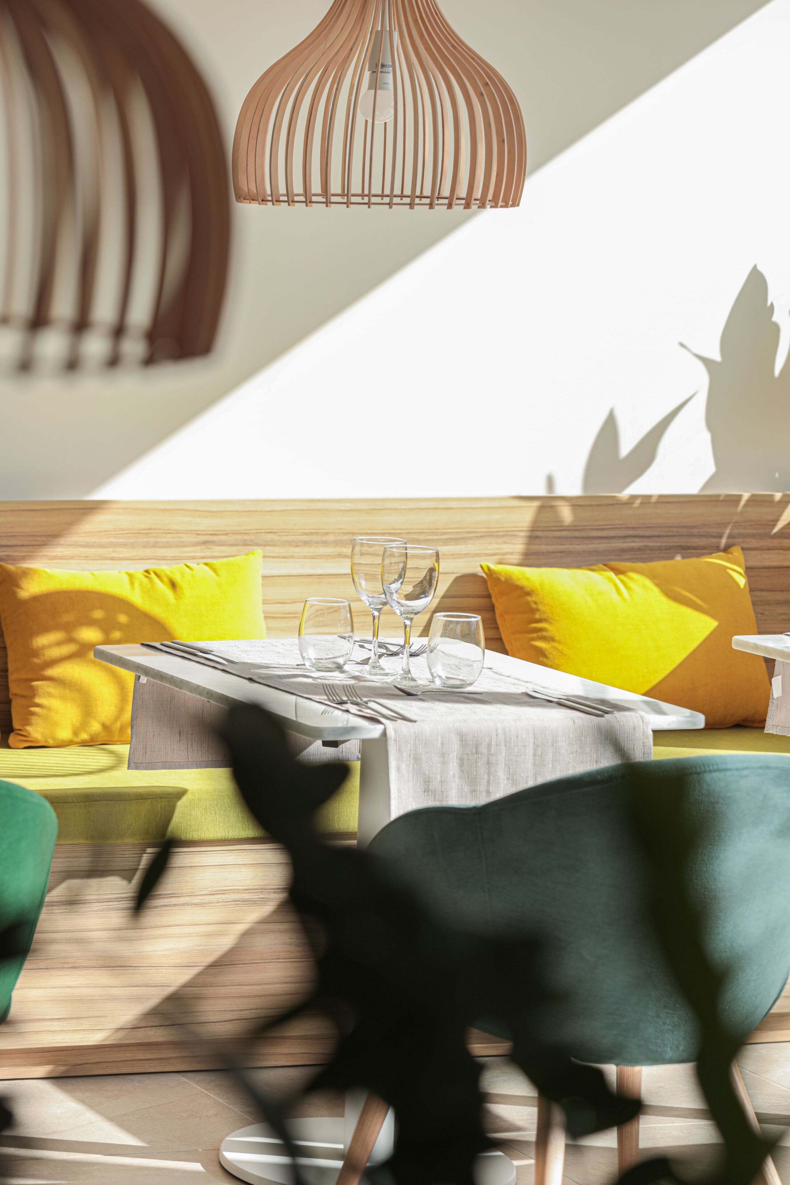 Un espace repas moderne avec une table dressée pour deux personnes, un banc en bois avec des coussins jaunes et une suspension lumineuse.