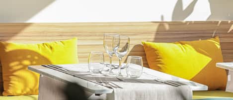 Un espace repas moderne avec une table dressée pour deux personnes, un banc en bois avec des coussins jaunes et une suspension lumineuse.