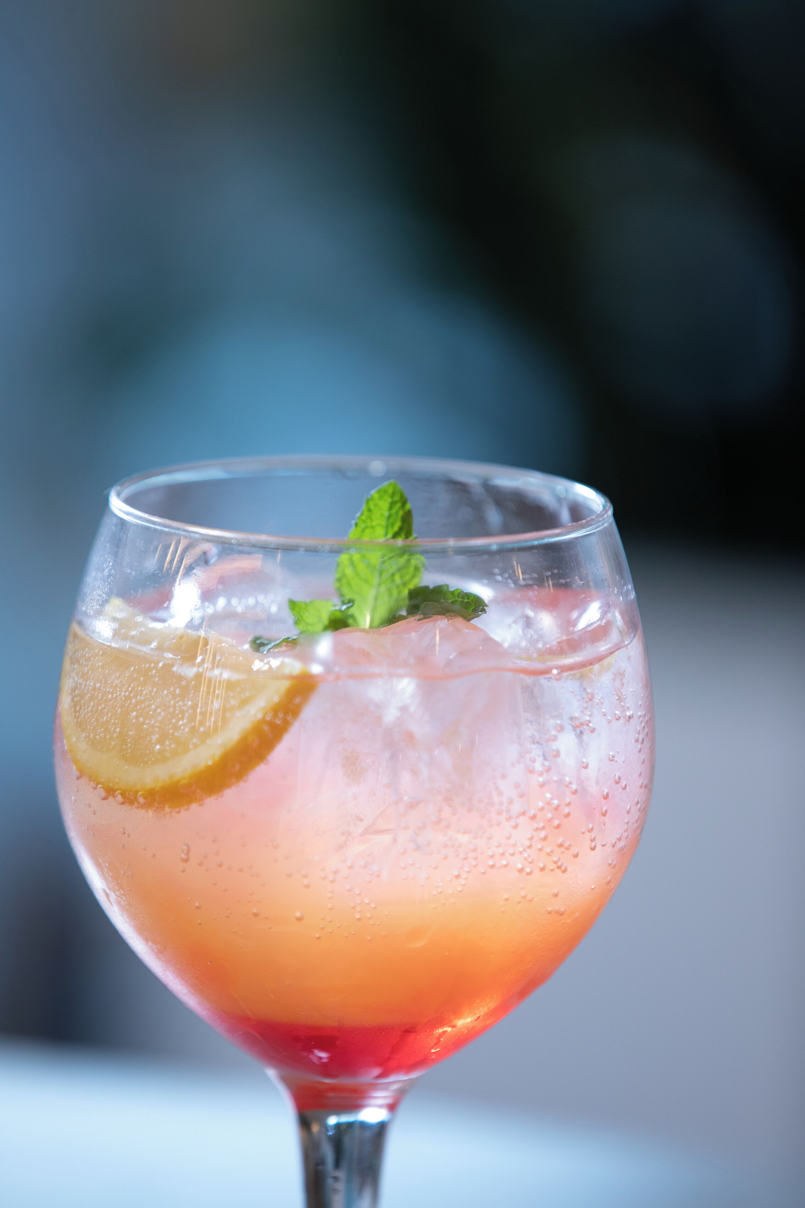 Un vaso transparente con una bebida rosada, hielo, una rodaja de naranja y una hoja de menta.