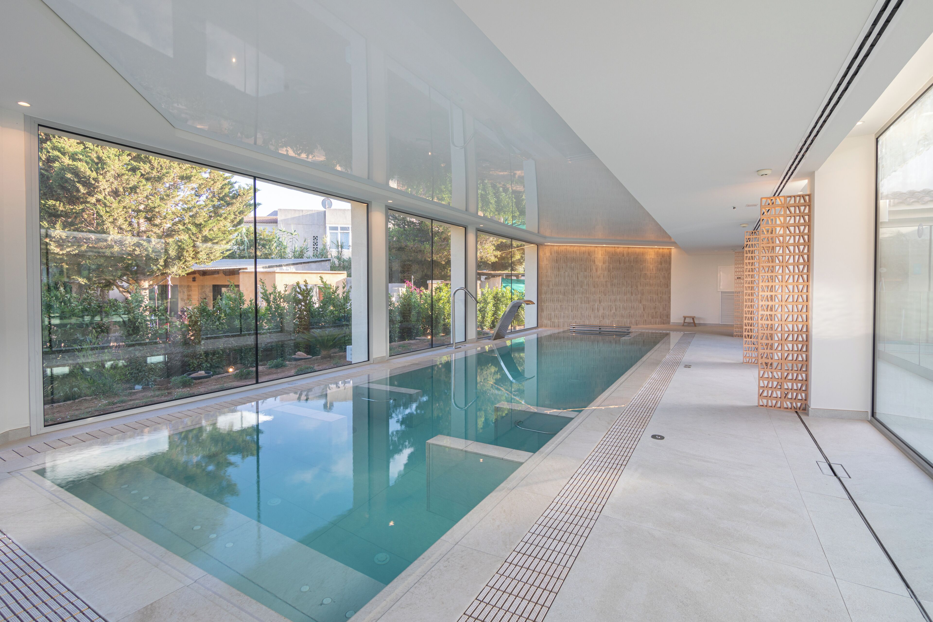 Une piscine intérieure moderne avec des parois en verre transparent, un sol carrelé et un panneau décoratif en bois sur le côté droit.