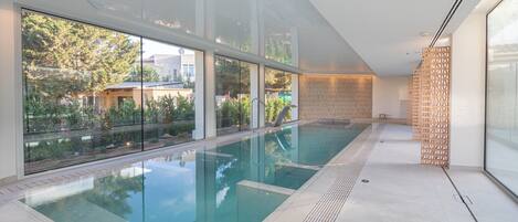 Une piscine intérieure moderne avec des parois en verre transparent, un sol carrelé et un panneau décoratif en bois sur le côté droit.