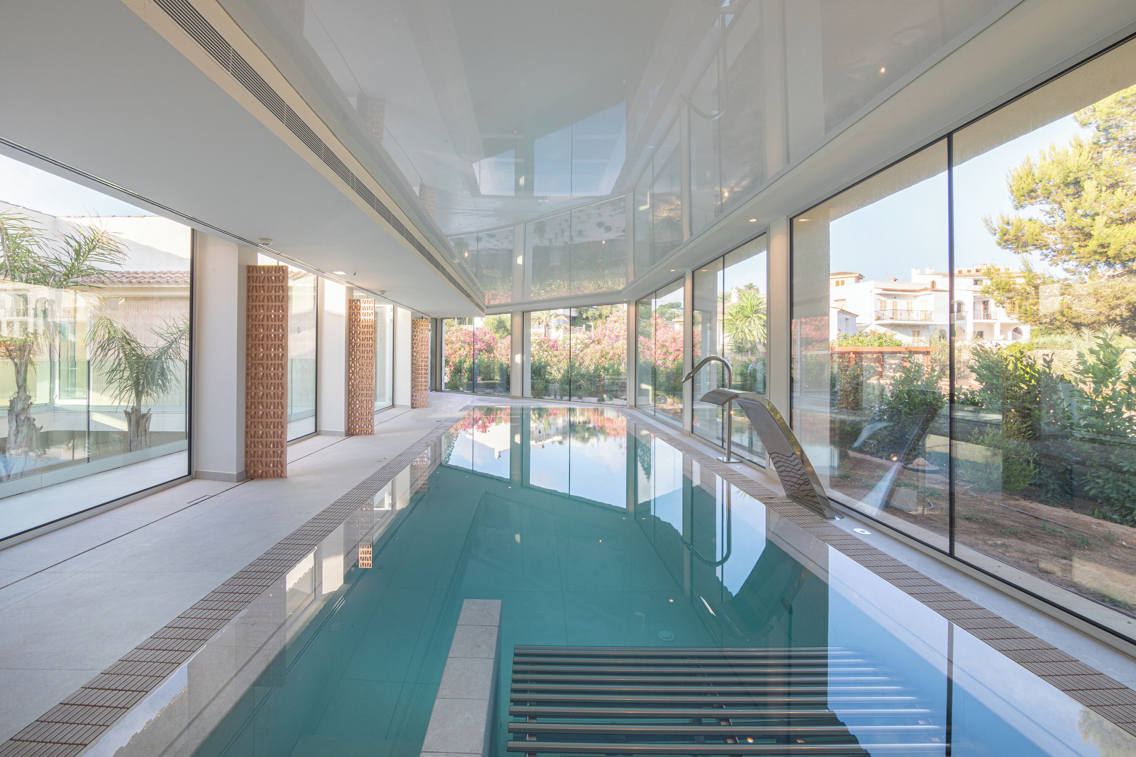 An indoor swimming pool with clear glass walls, a tiled floor, and a view of the surrounding garden and buildings.
