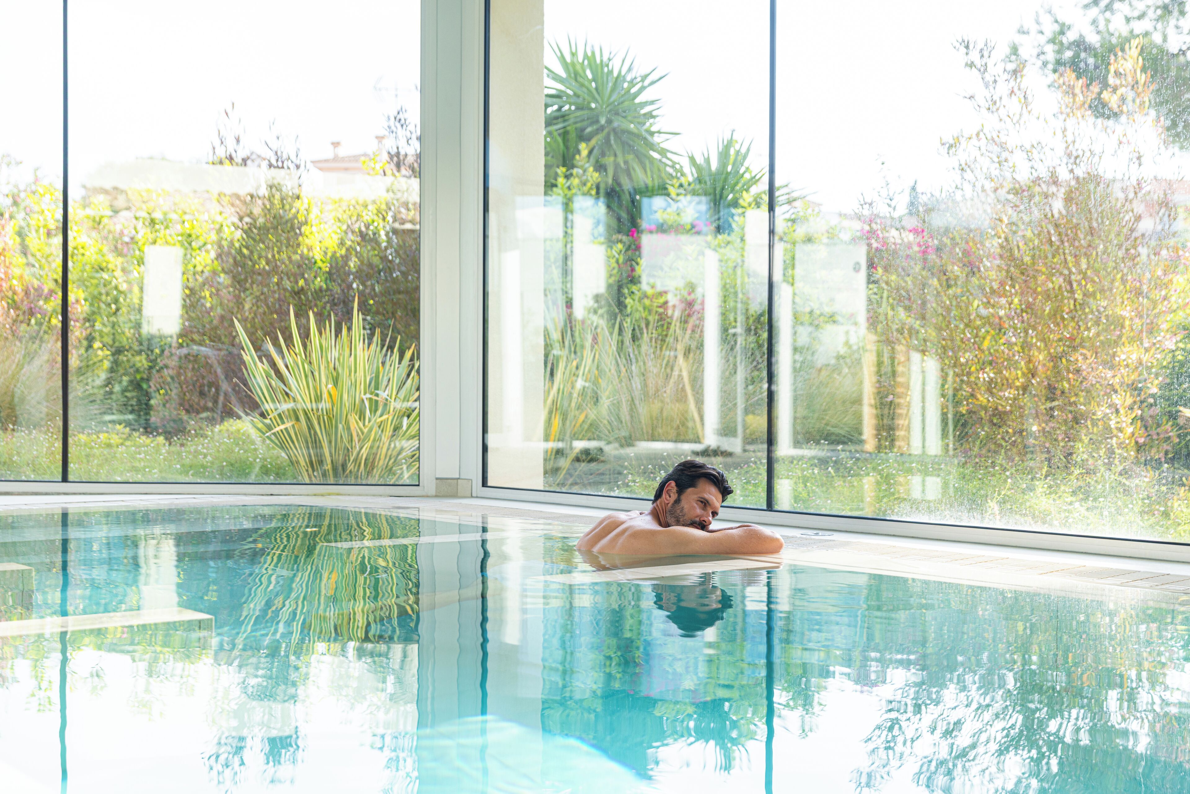 A man relaxing in a clear indoor swimming pool with large windows offering a view of lush greenery and a building in the background.