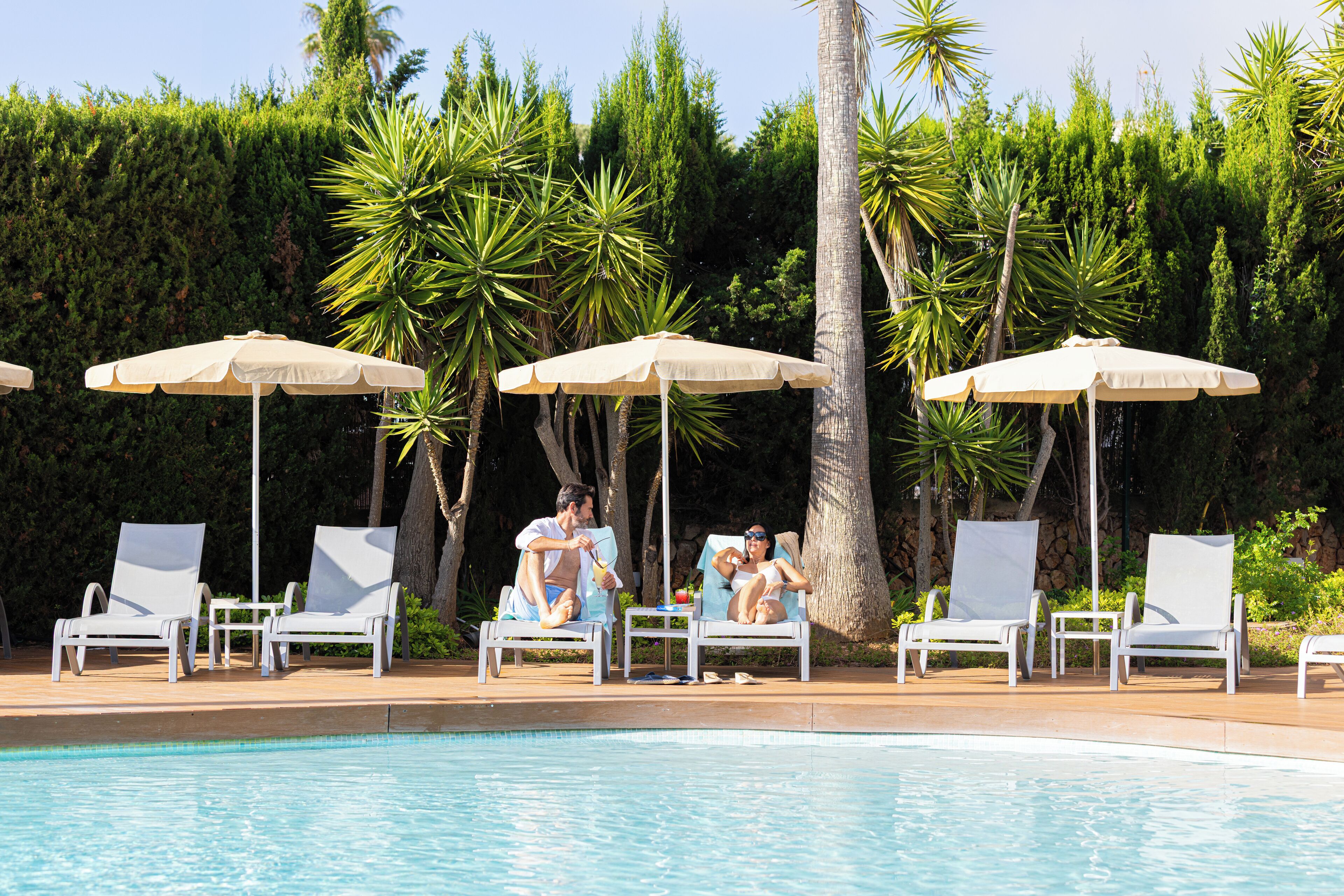 Un espace aménagé au bord de la piscine, avec des chaises longues et des parasols, entouré de palmiers et d’une haie.