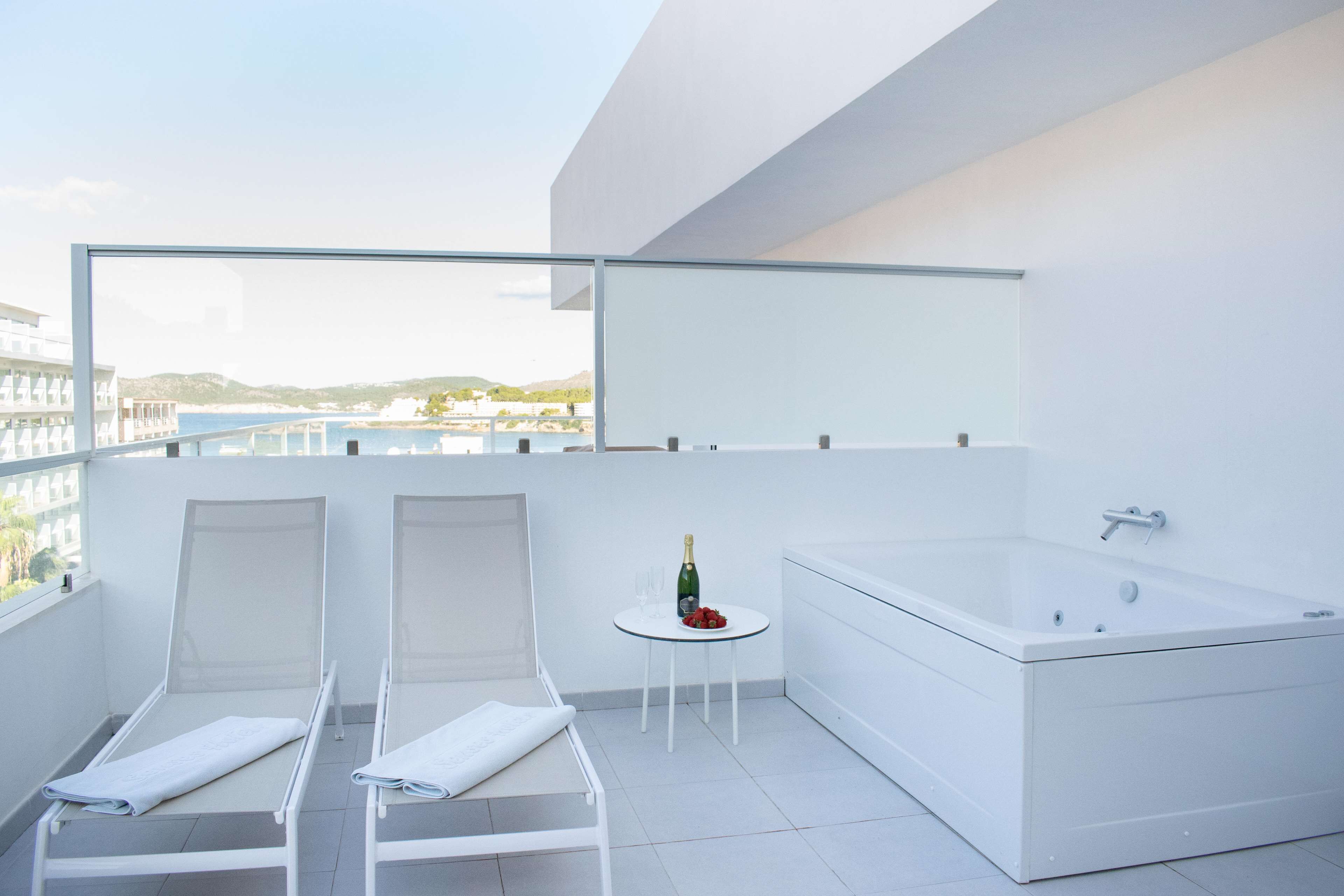 Ein moderner Balkon mit einer weißen Badewanne, zwei Liegestühlen und einem kleinen Tisch mit Flasche und Gläsern.