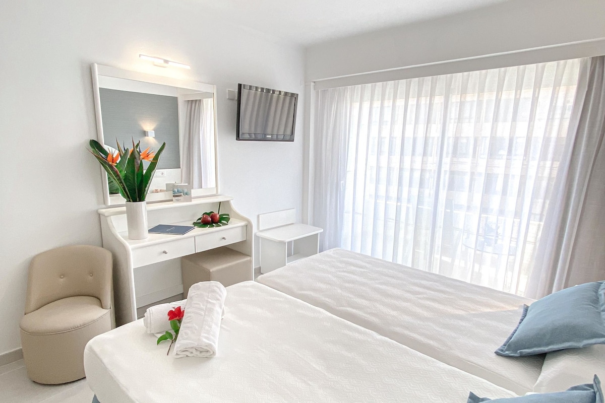 Une chambre d’hôtel avec un lit, un bureau, une chaise, un miroir et une fenêtre avec des rideaux.