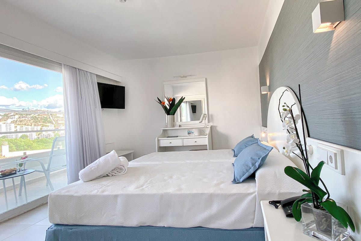 Una habitación de hotel moderna con una cama grande, un balcón con vistas, un escritorio con teléfono y un televisor montado en la pared.