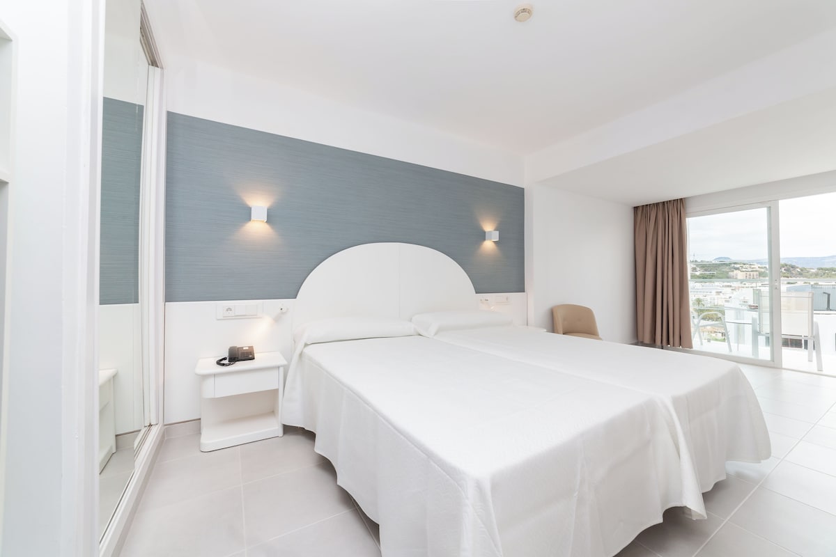 A modern hotel room with a large bed, a nightstand, a wall-mounted light, and a view of the city.