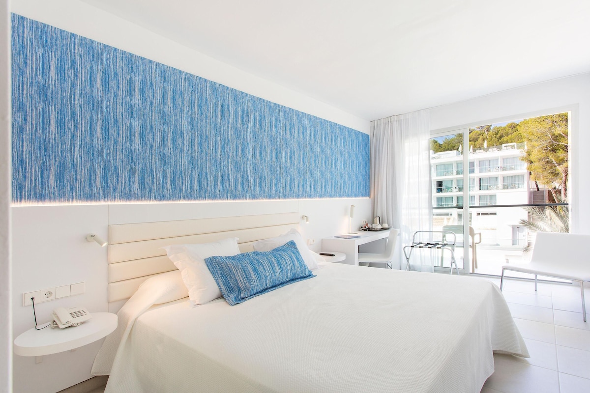 Une chambre d’hôtel avec un grand lit, une table de chevet avec un téléphone, un bureau avec une chaise et un balcon offrant une vue sur les bâtiments et les arbres.