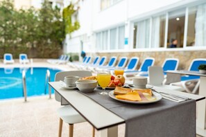 A breakfast setup with a poolside view, including a table with breakfast items, a glass of orange juice, and a view of the pool and lounge chairs.