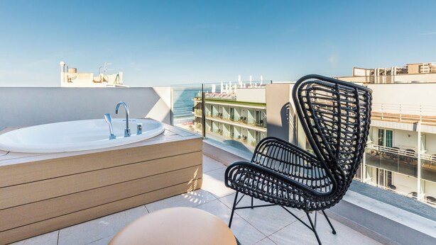 A balcony with a bathtub, a wicker chair, and a view of the city. - Hotel Cabot Playa Grande - Adults Only (Palma de Mallorca)