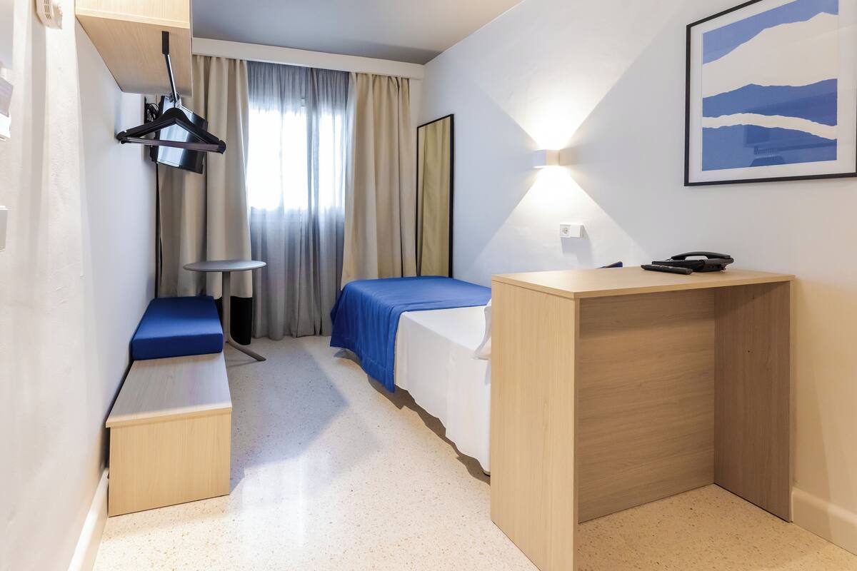 A hotel room with a bed, a desk, a small table, a bench, and a large window with curtains.