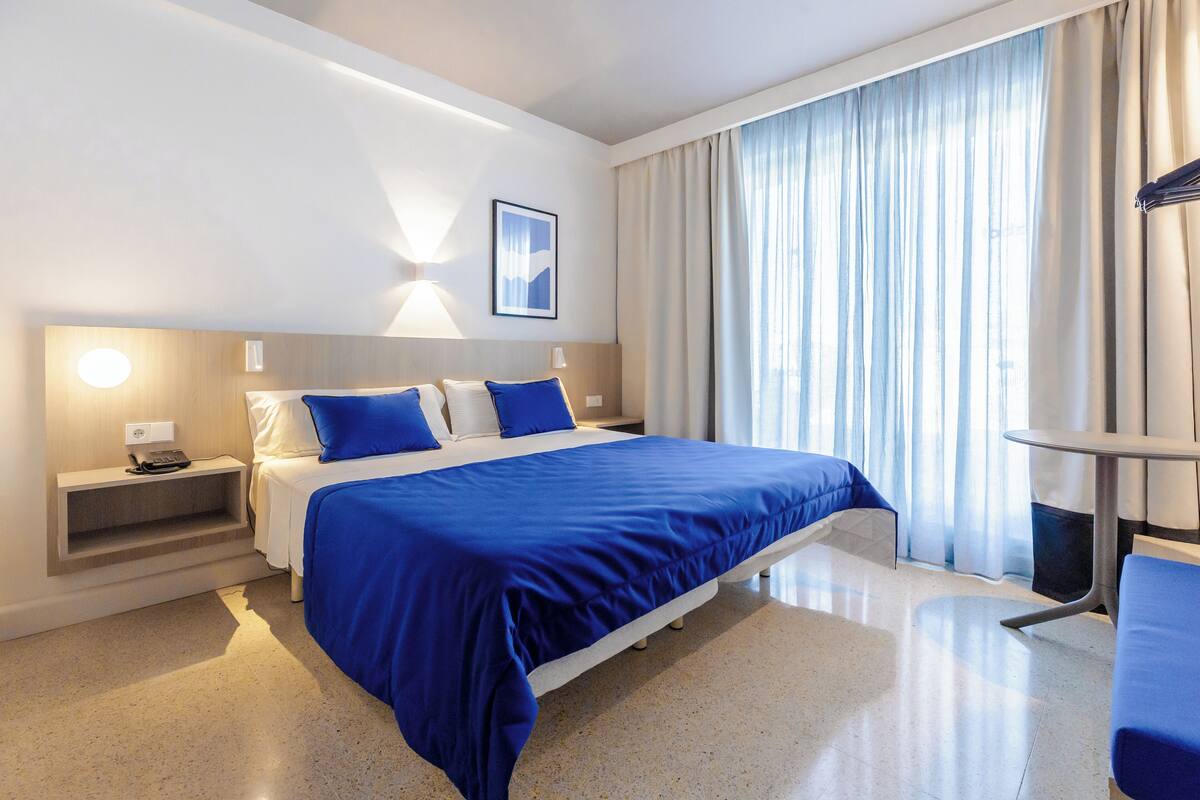 A modern hotel room with a large bed, a bedside table, a desk, and a chair.