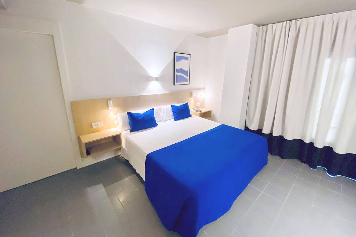 A hotel room with a bed, a nightstand, a wall-mounted lamp, and a window with curtains.