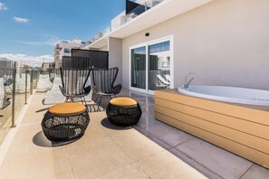 A modern outdoor balcony with a glass balustrade, two black wicker chairs with yellow cushions, a round wicker ottoman, a white bathtub, and a glass door leading to a balcony with white lounge chairs. - Hotel Cabot Playa Grande - Adults Only (Palma de Mallorca)