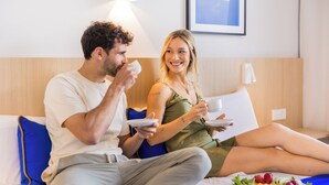 A couple sitting on a bed with a breakfast tray of fruit in a hotel room. - Hotel Cabot Playa Grande - Adults Only (Palma de Mallorca)