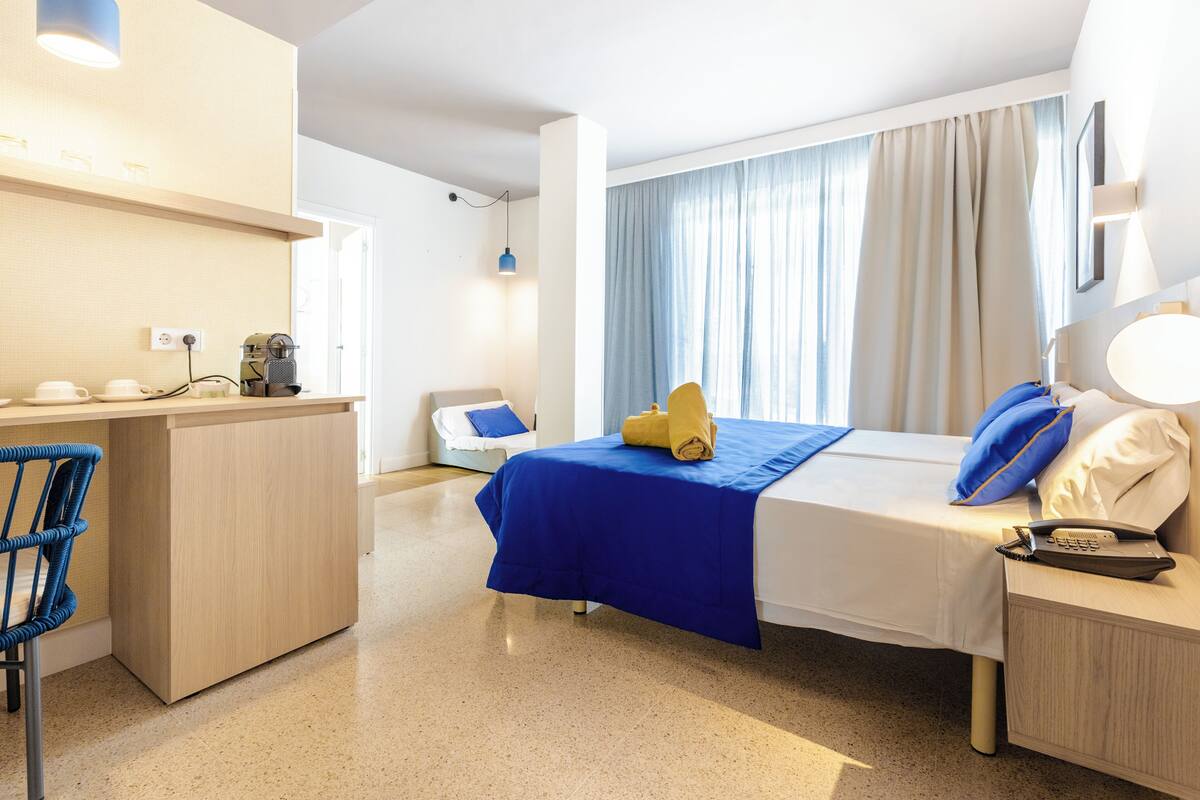 A hotel room with a bed, bedside table, desk, chair, and a window with curtains.
