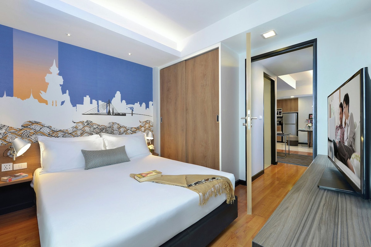 Ein modernes Hotelzimmer mit Bett, Fernseher, einer Stadtlandschaft als Wandgemälde und Blick in den Küchenbereich.