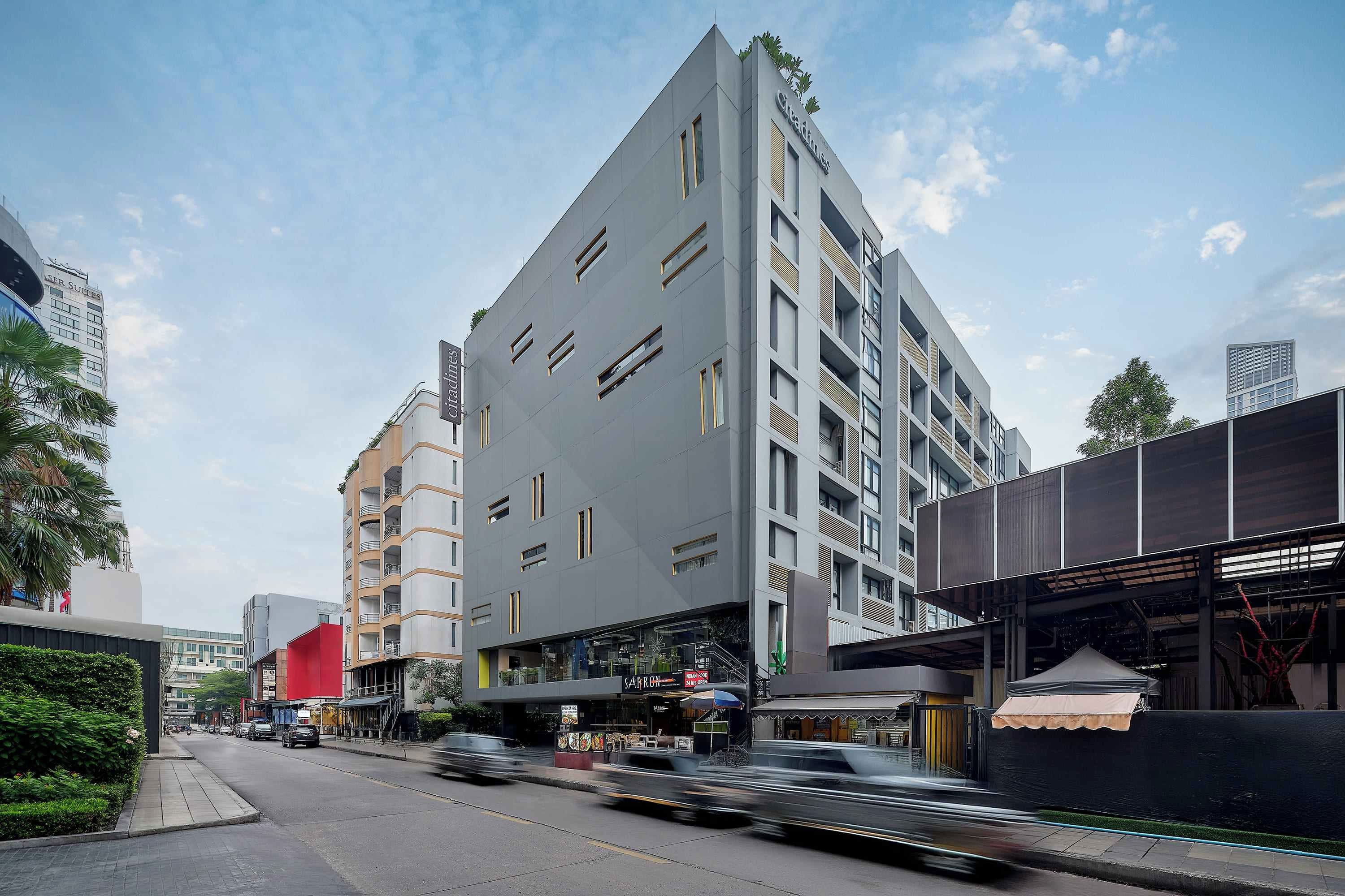 Un hôtel moderne, reconnaissable à son grand panneau « City Suites », doté de nombreux balcons et offrant une vue sur la rue, où l’on peut observer le passage des véhicules.
