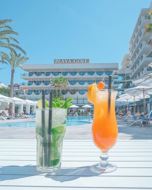 Dos cócteles sobre una mesa, con una piscina y un hotel llamado "PLAYA GOLF" al fondo.