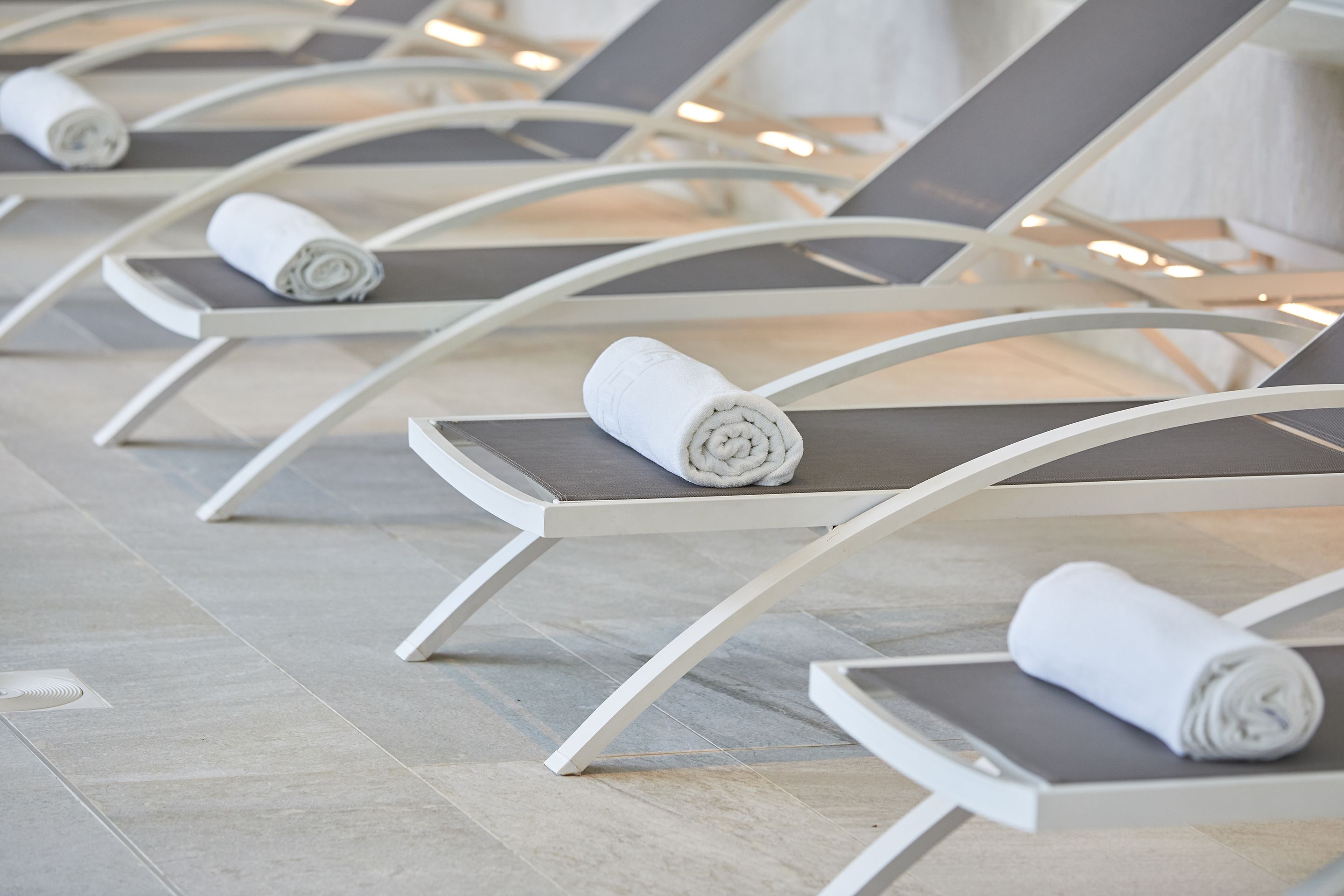 A row of white lounge chairs with grey cushions and rolled white towels placed on them.
