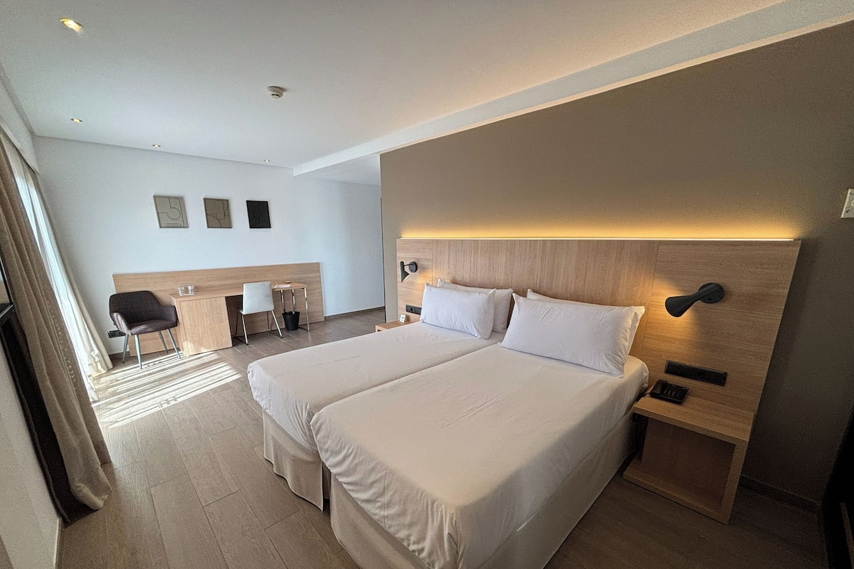 Une chambre d’hôtel avec deux lits, un bureau, une chaise, une petite table et une table de chevet.