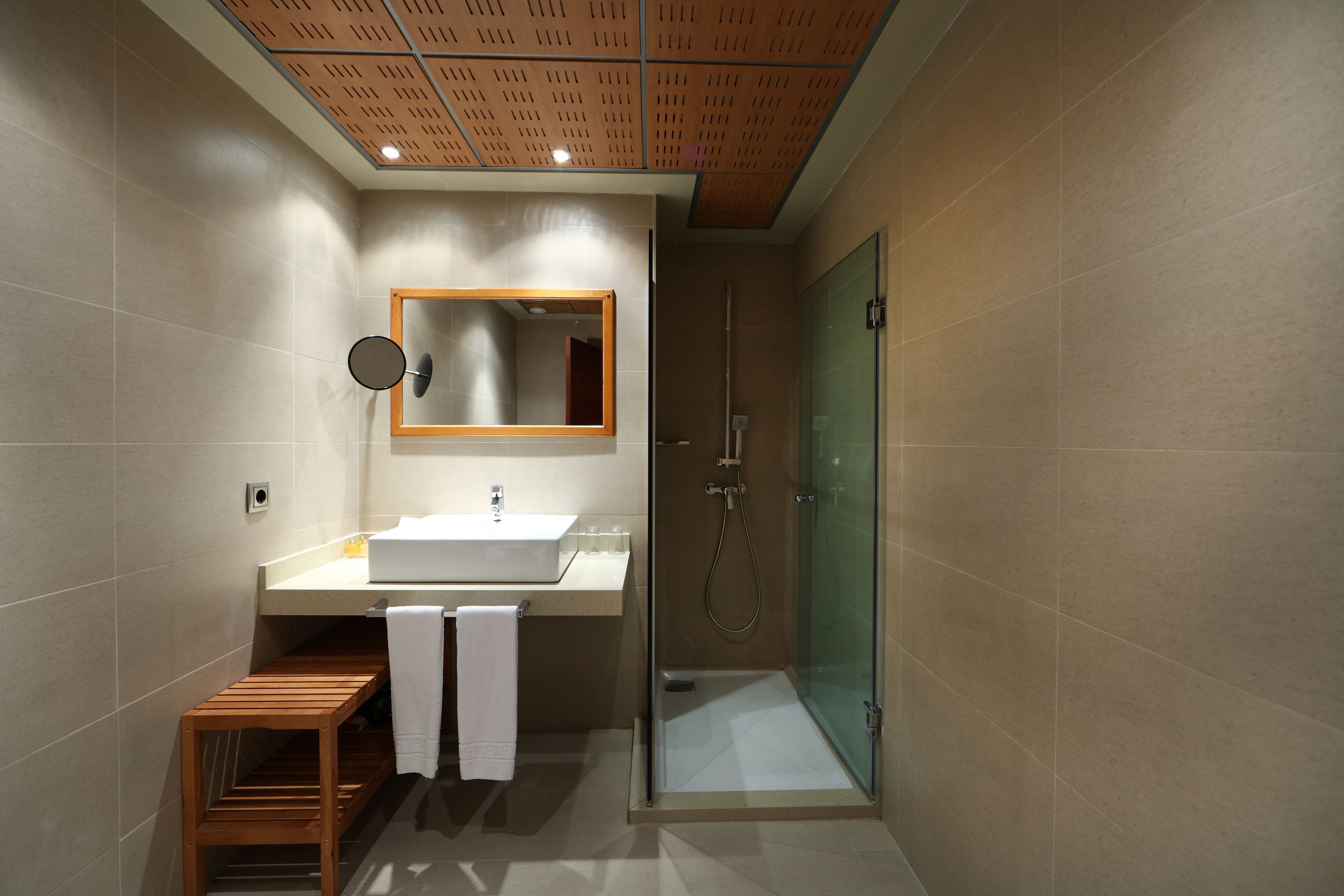 Un bagno moderno con una panca in legno, uno specchio grande, box doccia in vetro e lavandino bianco.