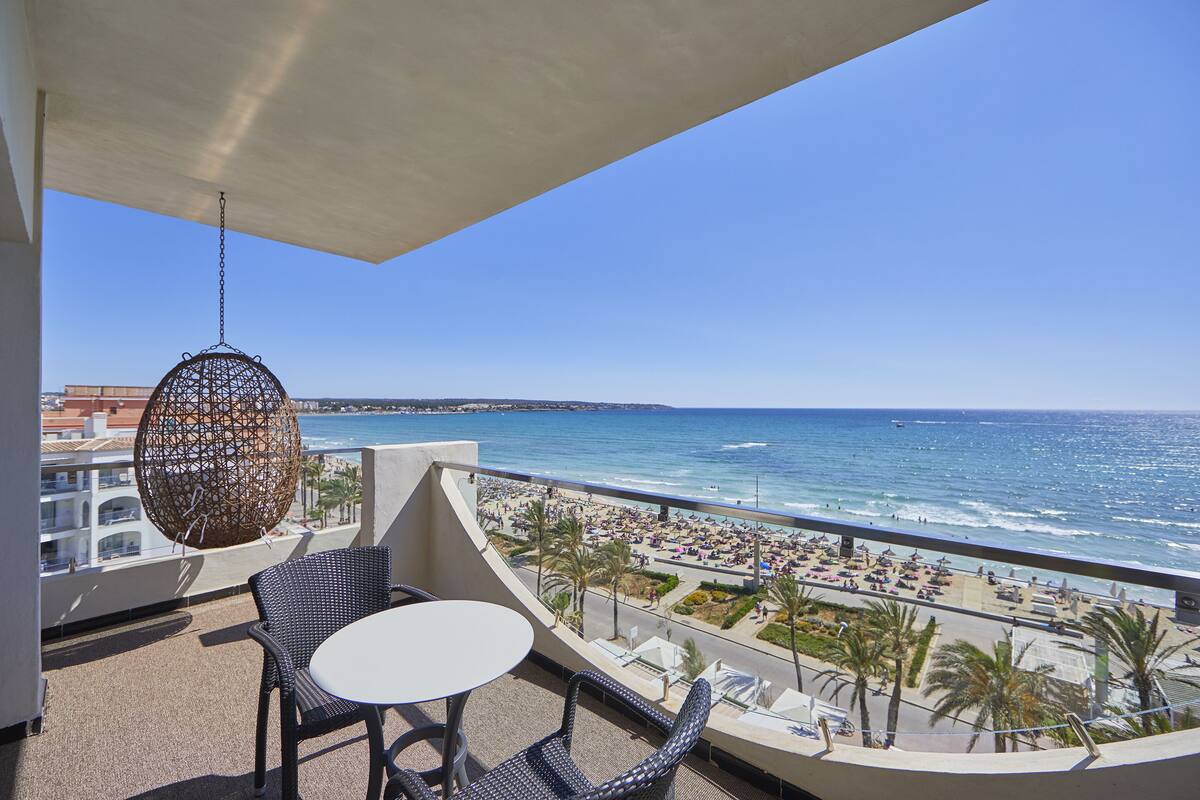 Un balcon avec des fauteuils en osier et une table, donnant sur une plage où l’on aperçoit des gens et des palmiers.