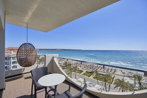 Un balcón con sillas de mimbre y una mesa, con vista a una playa con gente y palmeras.