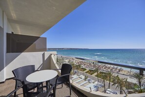 Un balcón con dos sillas de mimbre y una mesita, con vistas a una playa con sombrillas y gente.