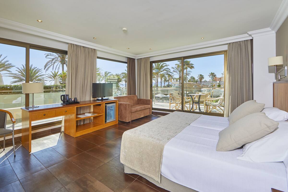 Une chambre d’hôtel avec un grand lit, un bureau avec une télévision, une chaise et un balcon donnant sur des palmiers et une piscine.