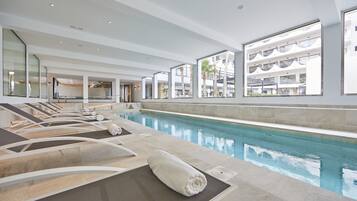 A modern indoor swimming pool with lounge chairs and a view of a multi-story building.