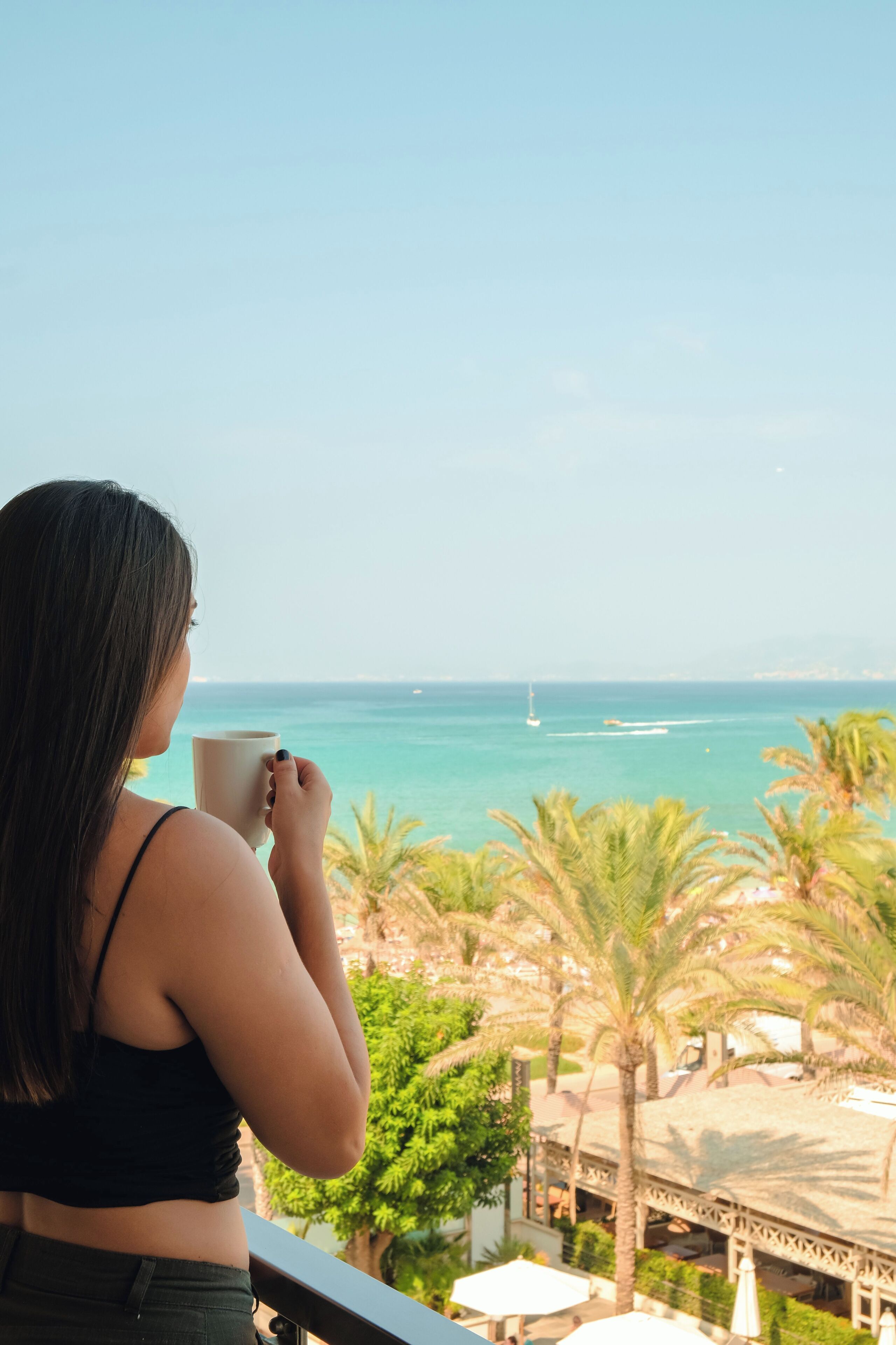 A person holding a mug, overlooking a beach with palm trees and a sailboat in the distance.