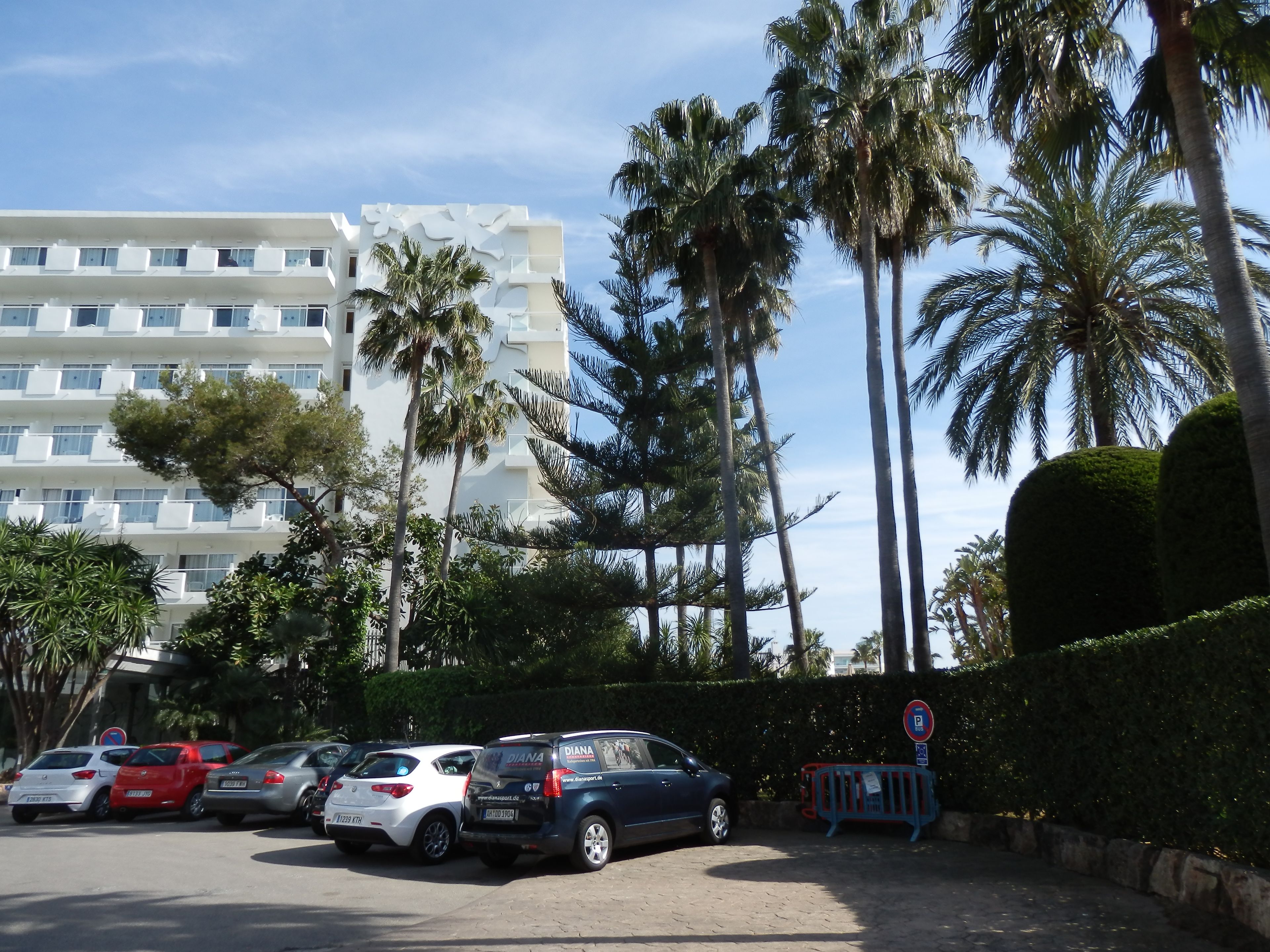 Un aparcamiento con varios coches, palmeras y un edificio con balcones.