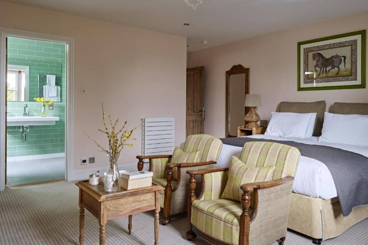 Une chambre d’hôtel avec un lit, deux fauteuils, une petite table avec un vase, un miroir et un tableau encadré au mur.