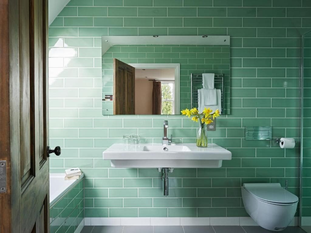 Un baño con azulejos verdes, un lavabo blanco, un espejo, un inodoro y un florero con flores amarillas.