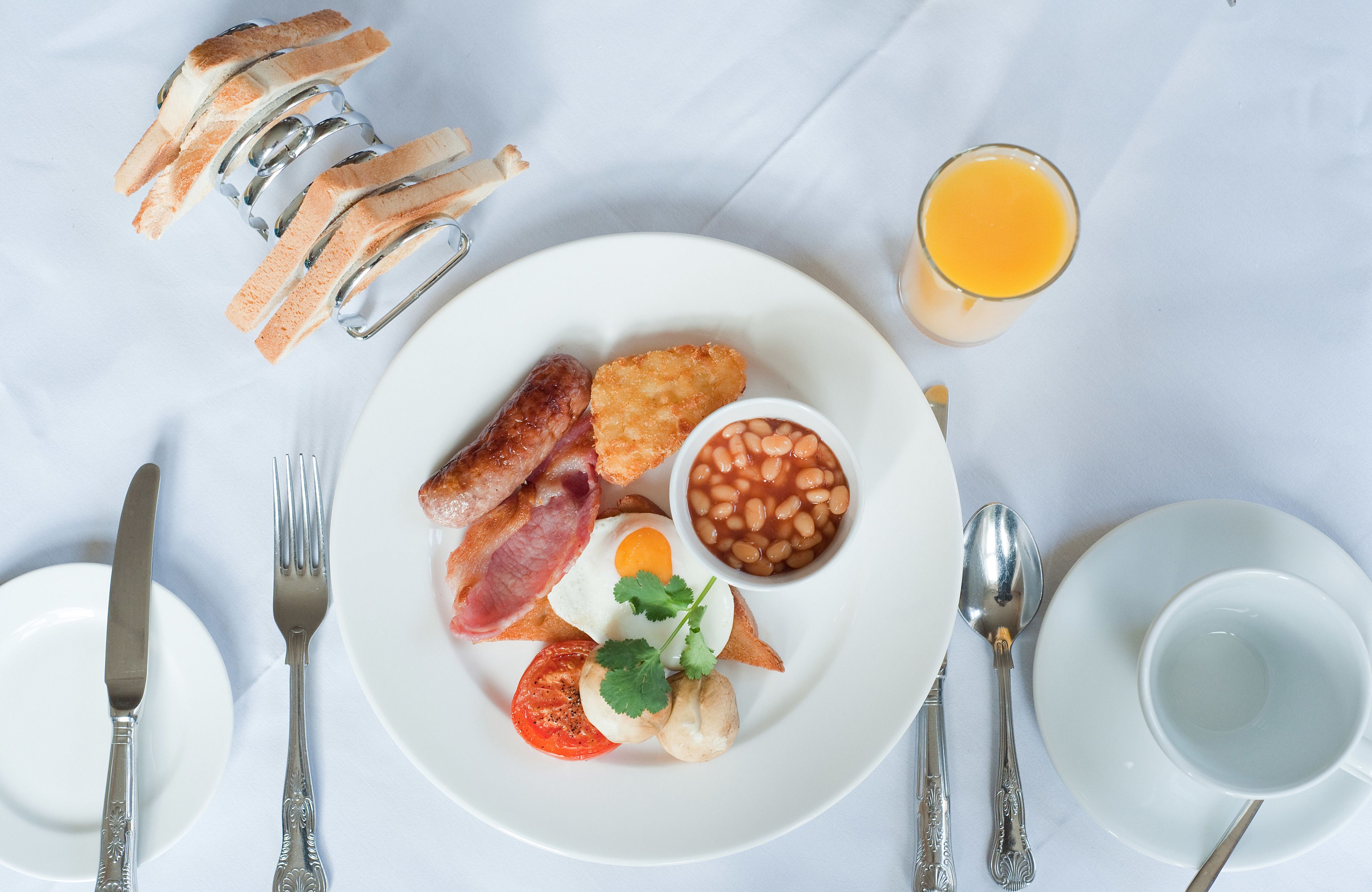 Ein Frühstücksteller mit Speck, Eiern, Bohnen und Toast, dazu ein Glas Orangensaft und Besteck auf einer weißen Tischdecke.