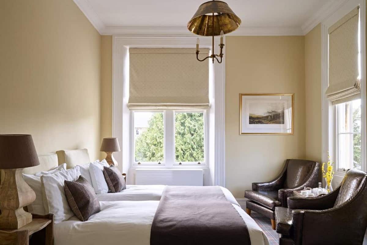 Une chambre d’hôtel avec deux lits, un coin salon avec un fauteuil, une lampe, un tableau au mur et une fenêtre donnant sur des arbres.