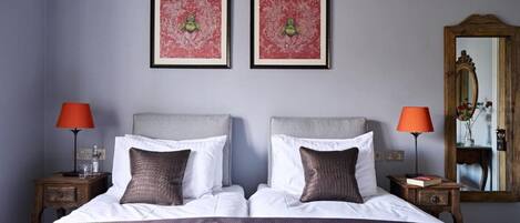 A double bed with white linens and two pillows, flanked by nightstands with lamps, in a room with a large mirror and two framed artworks on the wall.