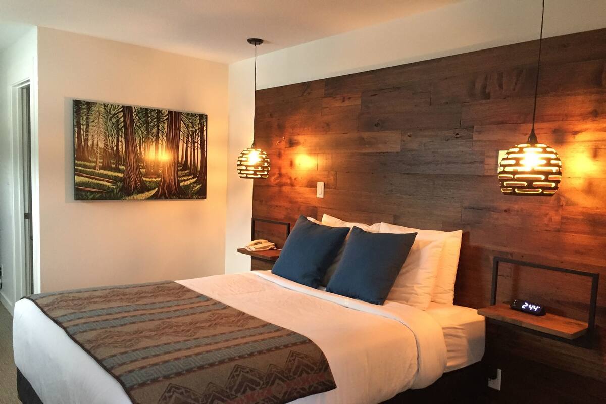 A neatly made bed with a patterned bedspread, two pillows, and a bedside table with a lamp. A painting of a forest scene hangs on the wall above the bed.