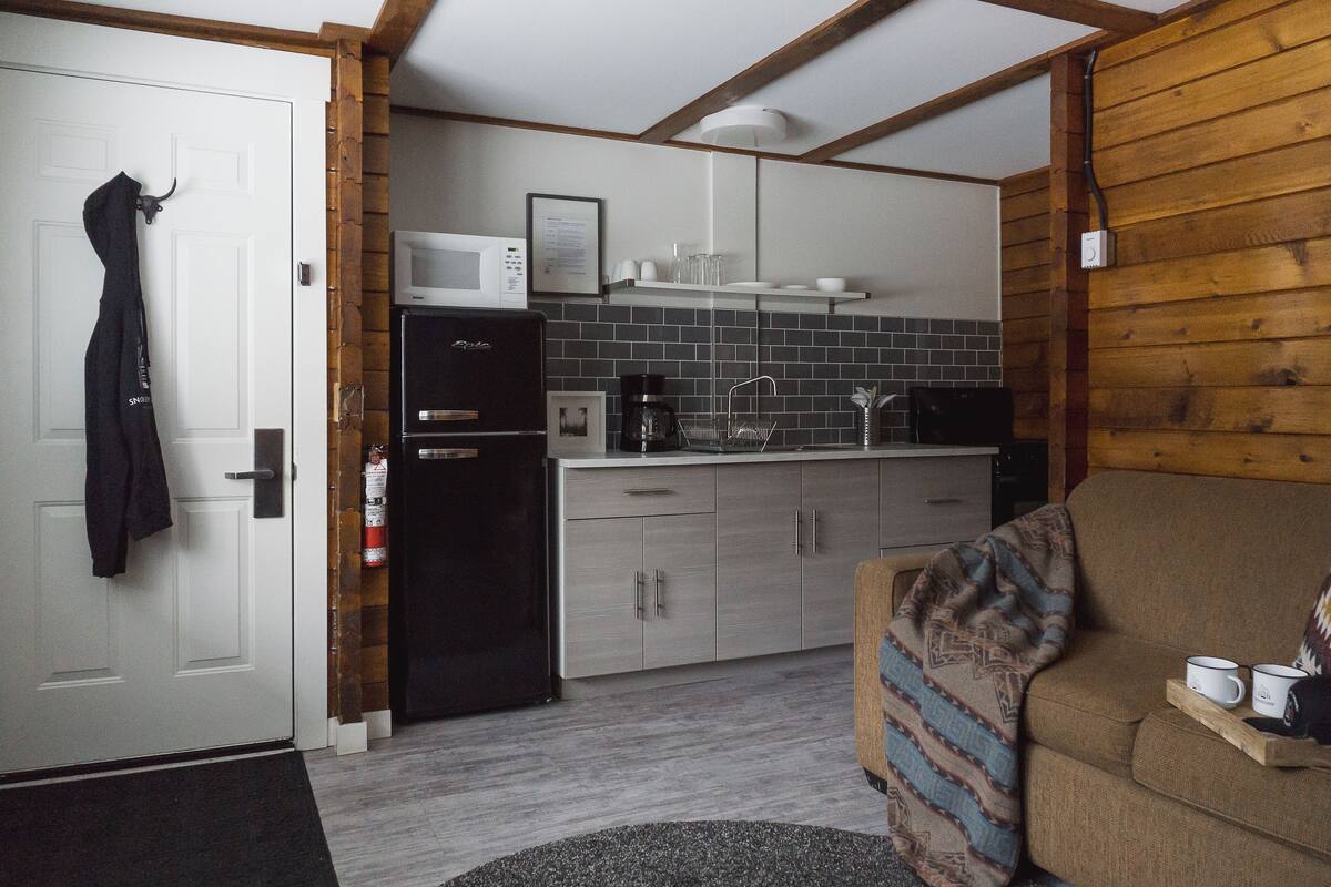 A compact kitchen with a microwave, refrigerator, and coffee maker. There is a wooden wall and a couch with a tray holding two mugs.