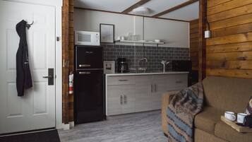 A compact kitchen with a microwave, refrigerator, and coffee maker. There is a wooden wall and a couch with a tray holding two mugs.
