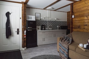 A compact kitchen with a microwave, refrigerator, and coffee maker. There is a wooden wall and a couch with a tray holding two mugs.
